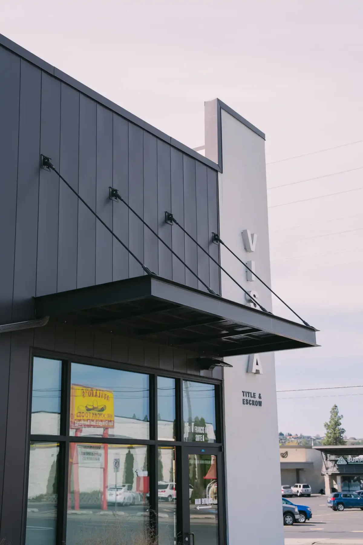 Mans Shop Entry Aluminum Awning The image shows a modern commercial building with Vestis black powder coated aluminum awning extending over the store’s front glass doors and entryway.