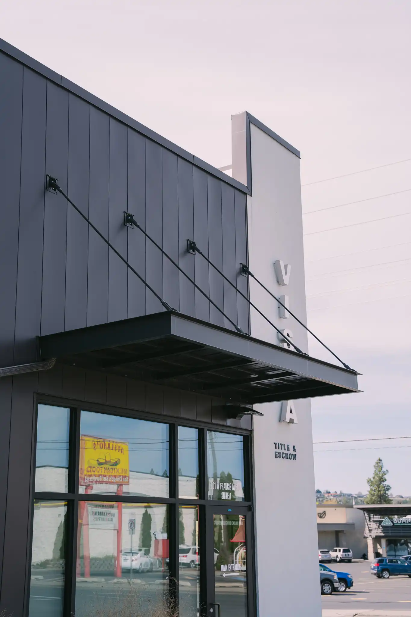 The image shows a modern commercial building with Vestis black powder coated aluminum awning extending over the store’s front glass doors and entryway.